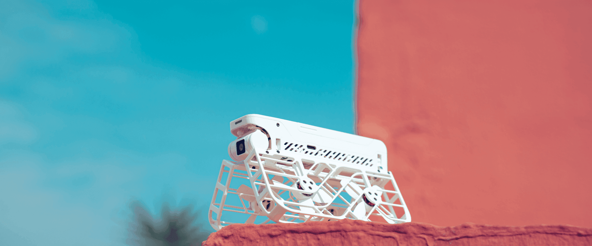 White drone with protective cage resting on an orange surface, against a sky-blue background.